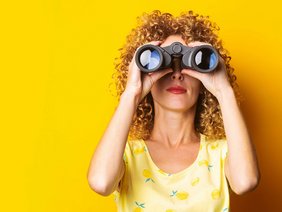 Frau mit Fernglas vor gelbem Hintergrund schaut zum Betrachter