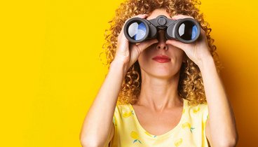 Frau mit Fernglas vor gelbem Hintergrund schaut zum Betrachter