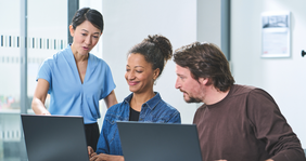 Zwei Mitarbeiter*innen sitzen vor Laptops, während eine dritte etwas erläutert | ©DVV